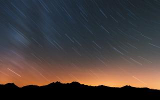 A mountain range is silhouetted by the orange dawn behind it. Most of the sky is still dark, and time-lapse trails of the paths of the stars show the spinning of the Earth.