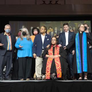 UUs leaders, clergy gathered on stage with a hand on each other and Pres-elect Sofia Betancourt seated at the center