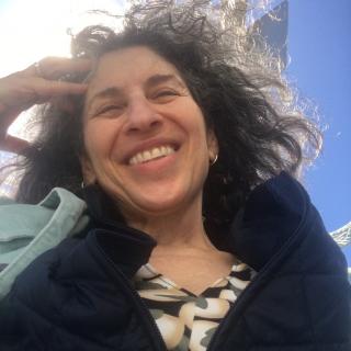 This is a shoulders-up photo of Susan Dana Lawrence, a white woman with salt and pepper wavy hair, smiling into the sun. She is wearing a print shirt, a black sweater, and light blue, collared jacket.